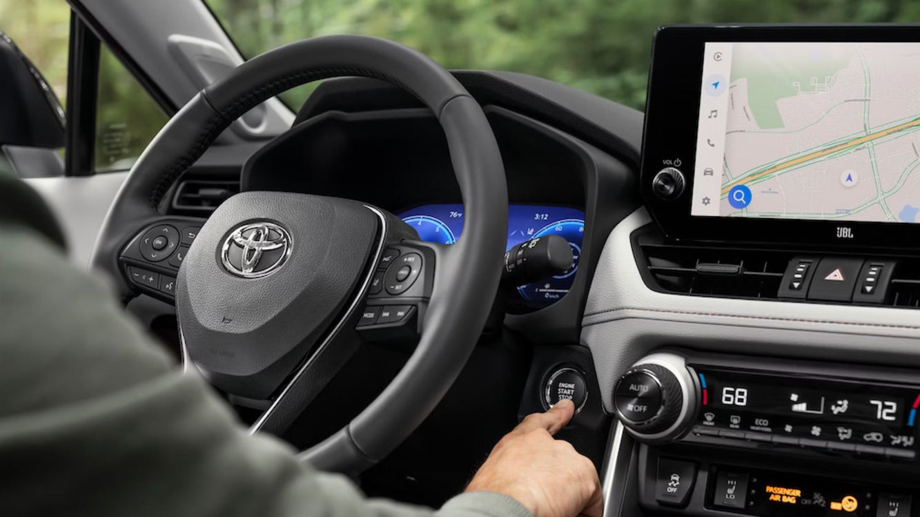 The steering wheel and console of a Toyota Prius, with a white person’s hand pressing the start button